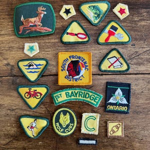 May include: A collection of embroidered scout patches in various shapes and colours, including a deer, a fish, a bicycle, a runner, a wolf head, and text reading South Frontenac District, 1st Bayridge, and Ontario with a white trillium flower.