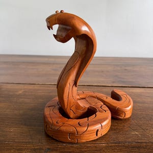 Peut inclure: Une sculpture en bois brun représentant un serpent cobra en position enroulée. La figurine est composée de plusieurs pièces imbriquées, avec une tête relevée, une bouche ouverte, des crocs visibles et deux petits yeux arrondis sur le dessus.