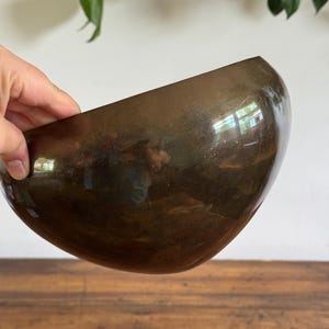 May include: A deep, rounded bowl with a smooth, dark brown metallic finish. The surface is highly reflective, showing light highlights and a glossy sheen across its curved exterior. The bowl has a simple, clean silhouette with no visible patterns.
