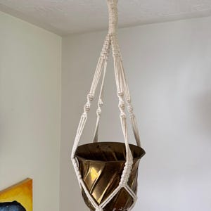 May include: A metallic bronze-coloured planter with a diagonal ribbed texture held by an off-white macrame cord hanger. The hanger features knotted patterns and terminates in a silver-toned metal ring at the top for ceiling suspension.