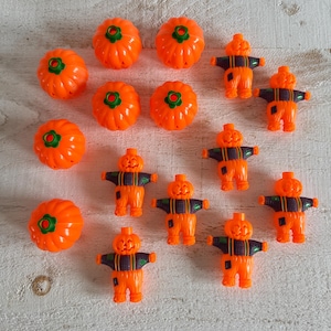 Könnte beinhalten: Eine Sammlung von orangefarbenen Halloween-Dekorationen aus Kunststoff, darunter kürbisförmige Ornamente und Vogelscheuchenfiguren. Die Kürbisse haben grüne Akzente, und die Vogelscheuchen tragen schwarz-violette Outfits. Die Artikel sind auf einer hellen Holzoberfläche angeordnet.