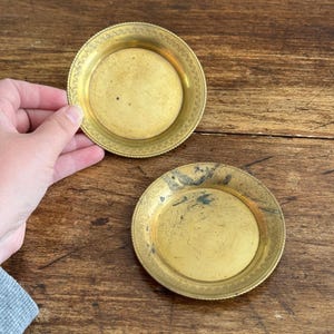 May include: Two circular brass trays with a smooth, flat centre and a decorative, textured border along the rim. The metal surfaces exhibit a golden hue with visible signs of age, including dark spots and patina on the lower tray.