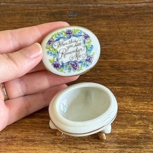May include: An oval porcelain trinket box with a removable lid featuring a floral wreath of purple and blue flowers. The text "When this you see Remember Me" is written in black script on the lid. The base has three small gold-toned feet.