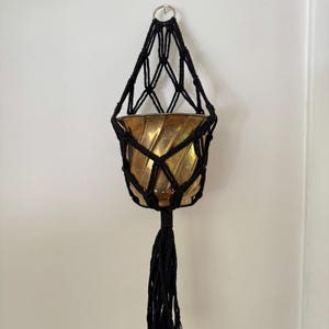 May include: A black macrame plant hanger featuring a knotted cord design and a long tassel at the bottom. The hanger holds a metallic gold-toned planter with a spiraled, ribbed texture. A silver metal ring is attached at the top for hanging.