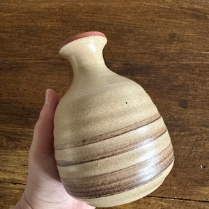 Peut inclure: Un vase en céramique beige avec des rayures horizontales marron. Le vase a un col étroit et un corps arrondi. Le bord est brun rougeâtre. Le vase a une esthétique rustique et terreuse.