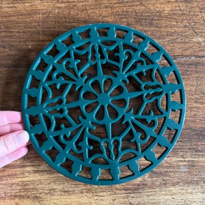 Peut inclure: Un dessous de plat circulaire en fonte vert foncé présentant un motif ajouré complexe avec un motif floral central entouré de volutes symétriques et d'une bordure géométrique, offrant une surface stable pour les ustensiles chauds.