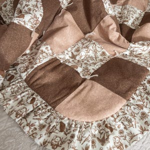May include: A brown and cream patchwork quilt with a ruffled edge. The quilt is made of squares of different fabrics, some with a floral pattern and some with a solid color. The quilt is folded in half and lying on a white surface.