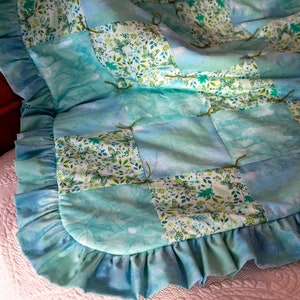 May include: A blue and green patchwork quilt with a ruffled edge. The quilt is made of squares of fabric with a floral pattern and butterflies.