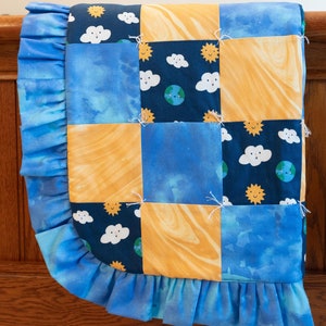 May include: A blue and yellow patchwork baby quilt with a ruffled edge. The quilt features a repeating pattern of clouds, suns, and the Earth.
