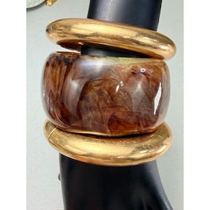 May include: A stack of three bracelets. The top and bottom bracelets are gold-colored, while the center bracelet is a marbled brown and cream color. The bracelets are displayed on a black mannequin hand.