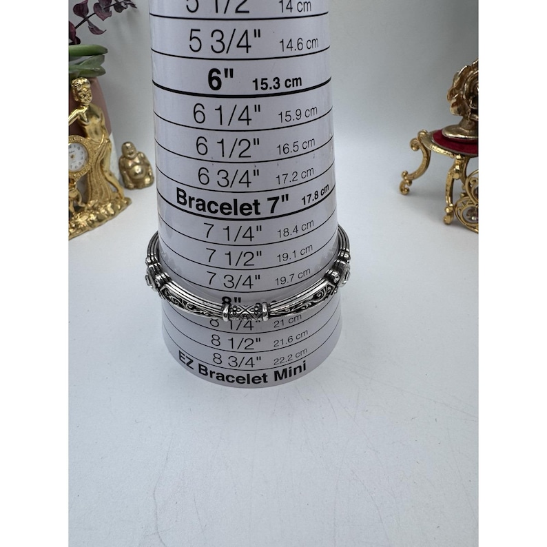 May include: A silver-toned bracelet with an ornate design is placed on a bracelet sizing tool. The tool measures up to 8 3/4 inches (22.2 cm). The bracelet is approximately 7 inches (17.8 cm) in diameter.