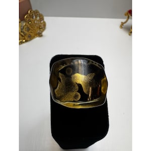 May include: A wide, black and gold cuff bracelet with a rectangular shape. The bracelet features gold leaf designs and is displayed on a black velvet stand. The cuff is a statement piece of jewelry.