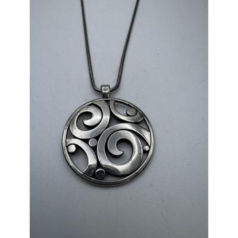 以下が含まれることがあります： 渦巻き模様と小さな円形のアクセントが特徴の、シルバーカラーのペンダントネックレス。ペンダントは、お揃いのシルバーチェーンから吊り下げられており、シンプルながらもエレガントなアクセサリーです。ペンダントの直径は約4cmです。