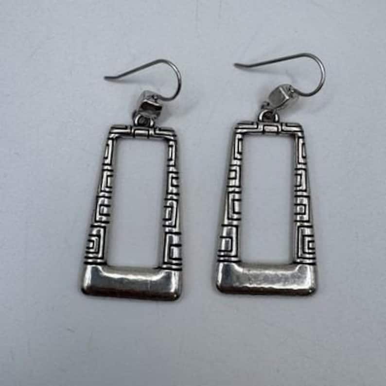 May include: A pair of silver-toned rectangular earrings. Each earring features a geometric design along the edges and a curved bottom. The earrings have a hook closure for pierced ears.