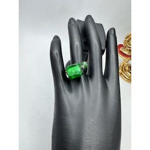 May include: A ring featuring a rectangular, vibrant green glass stone with a textured, speckled surface. The green glass is set in a silver-toned metal band that wraps around the finger, showcasing a clear, faceted accent piece at the top.