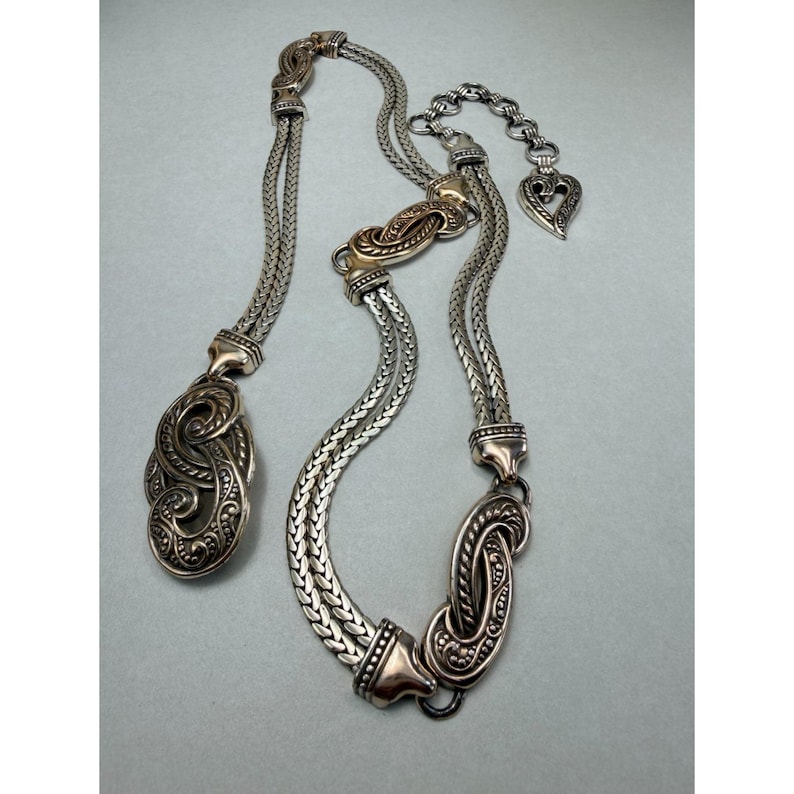 May include: A silver-toned necklace with an ornate pendant and chain. The necklace features a braided chain design with decorative clasps and a heart-shaped charm. The pendant has a detailed, swirling pattern.