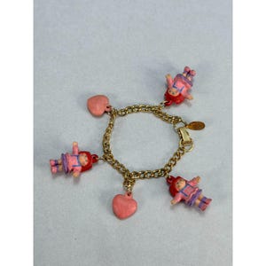May include: A gold-tone metal chain link charm bracelet featuring three small doll charms with red hair and pink outfits, alternating with two smooth pink heart-shaped charms. The bracelet includes a gold-tone metal clasp and a small circular tag.