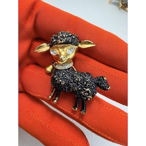 May include: A gold-toned sheep brooch with a black body and a gold head. The sheep has large, sparkling crystal eyes and a crystal-studded collar. The brooch is held against a bright orange background.