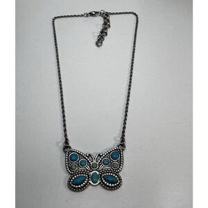 May include: A silver-toned necklace featuring a butterfly pendant. The butterfly pendant is adorned with turquoise and green-colored stones. The necklace has a chain with an adjustable clasp. The jewelry is displayed on a white surface.