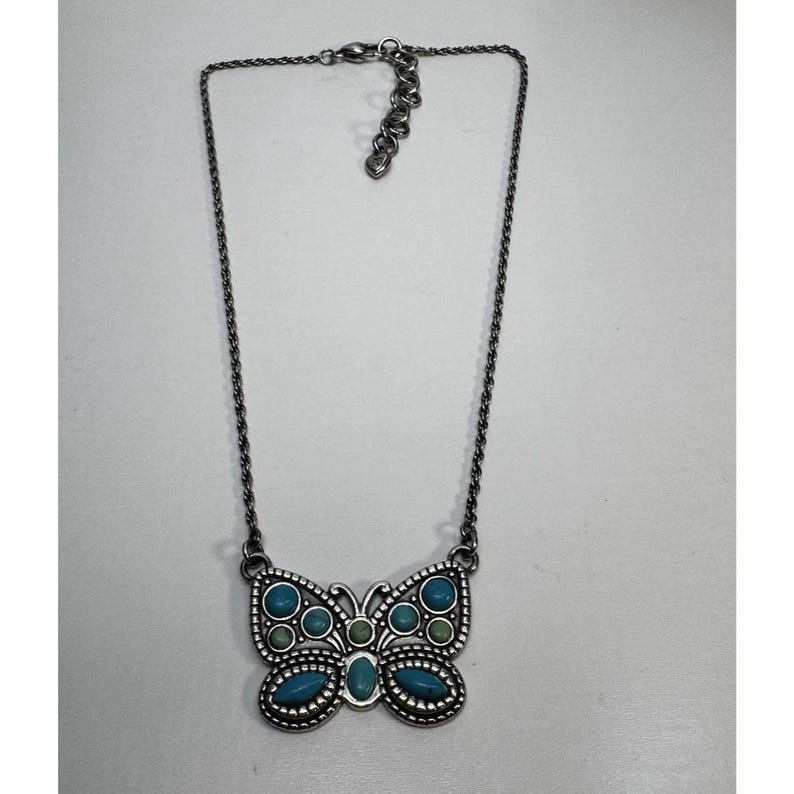 May include: A silver-toned necklace featuring a butterfly pendant. The butterfly pendant is adorned with turquoise and green-colored stones. The necklace has a chain with an adjustable clasp. The jewelry is displayed on a white surface.