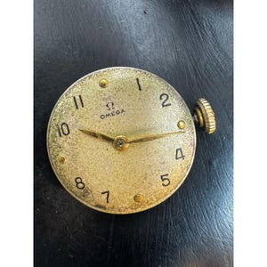 May include: A vintage Omega watch face with a gold-toned dial. The face features black numerals, gold hands, and the Omega logo. A gold crown is visible on the right side. The watch face has a textured, aged appearance.