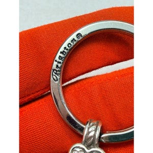 Peut inclure: Gros plan d'un porte-cl&eacute;s argent&eacute; avec le mot "Brighton" grav&eacute; sur l'anneau. Le porte-cl&eacute;s a un charm en forme de c&oelig;ur d&eacute;coratif avec un motif textur&eacute;. Le fond est un tissu orange vif.