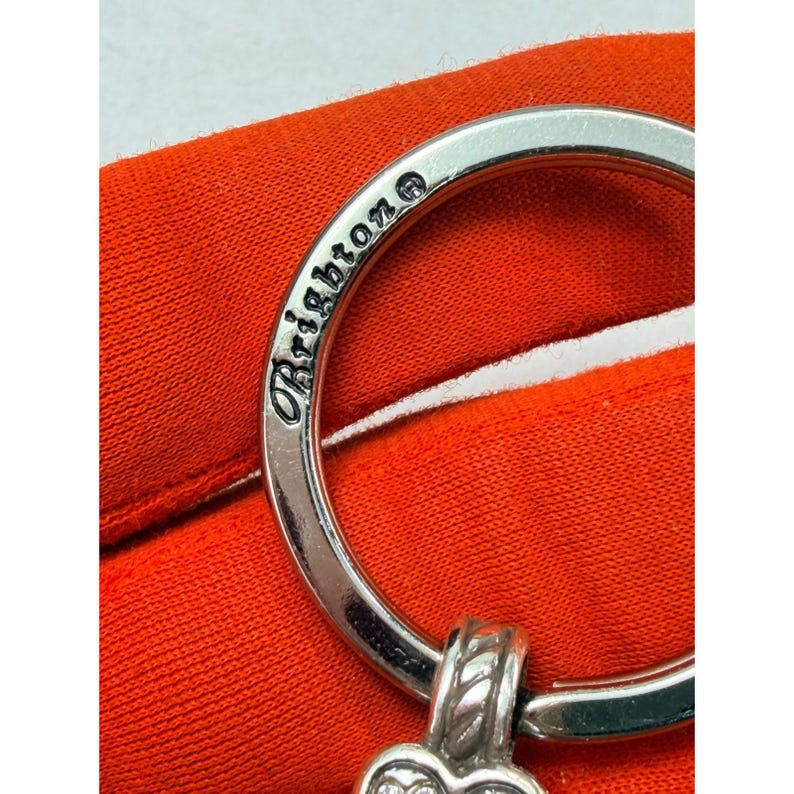 Peut inclure: Gros plan d'un porte-cl&eacute;s argent&eacute; avec le mot "Brighton" grav&eacute; sur l'anneau. Le porte-cl&eacute;s a un charm en forme de c&oelig;ur d&eacute;coratif avec un motif textur&eacute;. Le fond est un tissu orange vif.