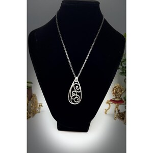 May include: A silver-toned pendant necklace with a teardrop-shaped openwork design. The pendant features swirling, scroll-like patterns and hangs from a delicate chain. The necklace is displayed on a black mannequin, showcasing its elegant design.
