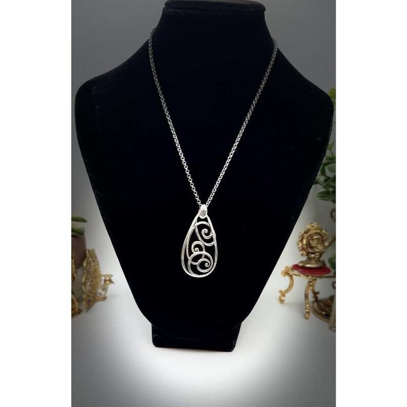 May include: A silver-toned pendant necklace with a teardrop-shaped openwork design. The pendant features swirling, scroll-like patterns and hangs from a delicate chain. The necklace is displayed on a black mannequin, showcasing its elegant design.