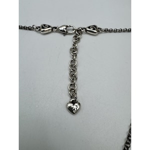 May include: Close-up of a silver-toned necklace clasp and chain. The necklace features a heart-shaped charm with a stylized design. The chain is composed of small, interconnected links, and the clasp is a lobster claw style.