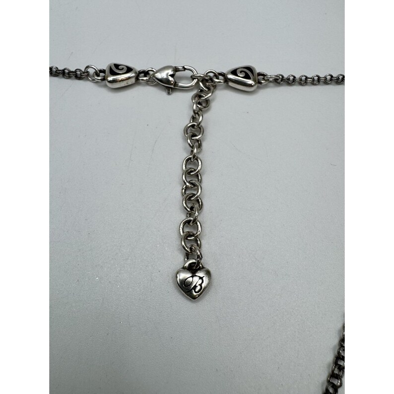 May include: Close-up of a silver-toned necklace clasp and chain. The necklace features a heart-shaped charm with a stylized design. The chain is composed of small, interconnected links, and the clasp is a lobster claw style.