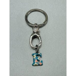 Peut inclure: Porte-cl&eacute;s argent&eacute; avec un charm en forme de lettre "E" &agrave; motifs floraux. Le porte-cl&eacute;s comporte un accent en forme de c&oelig;ur avec des cristaux transparents et un anneau porte-cl&eacute;s rond. La lettre "E" est peinte en bleu sarcelle avec des d&eacute;tails floraux roses et blancs.