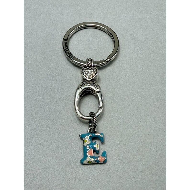 Peut inclure: Porte-cl&eacute;s argent&eacute; avec un charm en forme de lettre "E" &agrave; motifs floraux. Le porte-cl&eacute;s comporte un accent en forme de c&oelig;ur avec des cristaux transparents et un anneau porte-cl&eacute;s rond. La lettre "E" est peinte en bleu sarcelle avec des d&eacute;tails floraux roses et blancs.