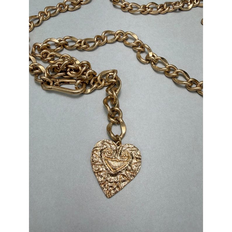 Puede incluir: Cintur&oacute;n de cadena dorado con un colgante de coraz&oacute;n texturizado. El cintur&oacute;n presenta una cadena de eslabones grandes y un colgante decorativo en forma de coraz&oacute;n con una superficie detallada. El cierre es un cierre simple de gancho y ojo.