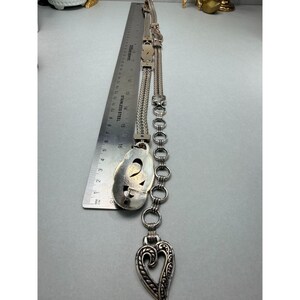 May include: A silver-toned necklace with a heart-shaped pendant and an oval-shaped pendant. The necklace is measured with a ruler. The heart pendant has a decorative openwork design. The necklace is made of metal.