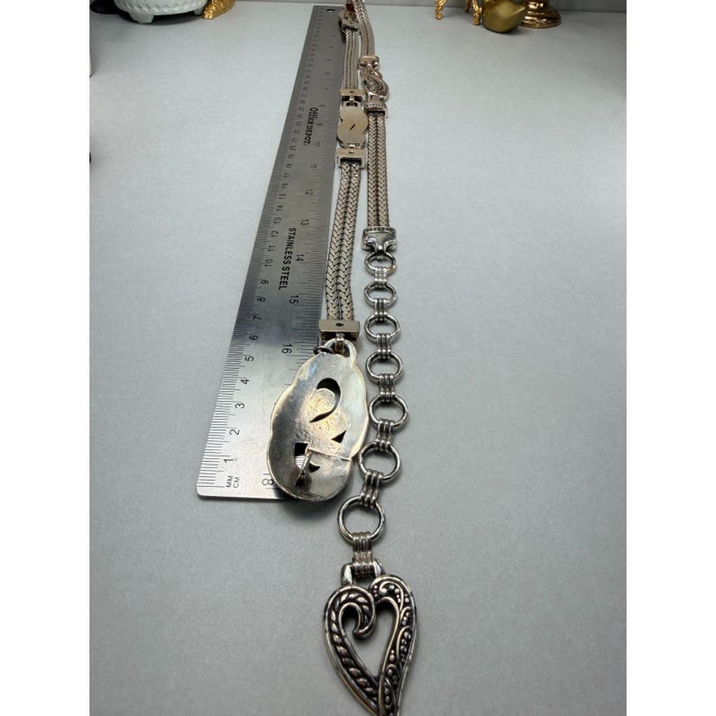 May include: A silver-toned necklace with a heart-shaped pendant and an oval-shaped pendant. The necklace is measured with a ruler. The heart pendant has a decorative openwork design. The necklace is made of metal.