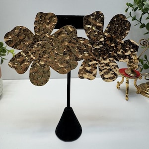 May include: Gold-toned, hammered metal flower earrings displayed on a black velvet jewelry stand. The earrings have a textured surface and a multi-toned gold and brown color scheme. The floral design is prominent, with each earring featuring multiple petals.
