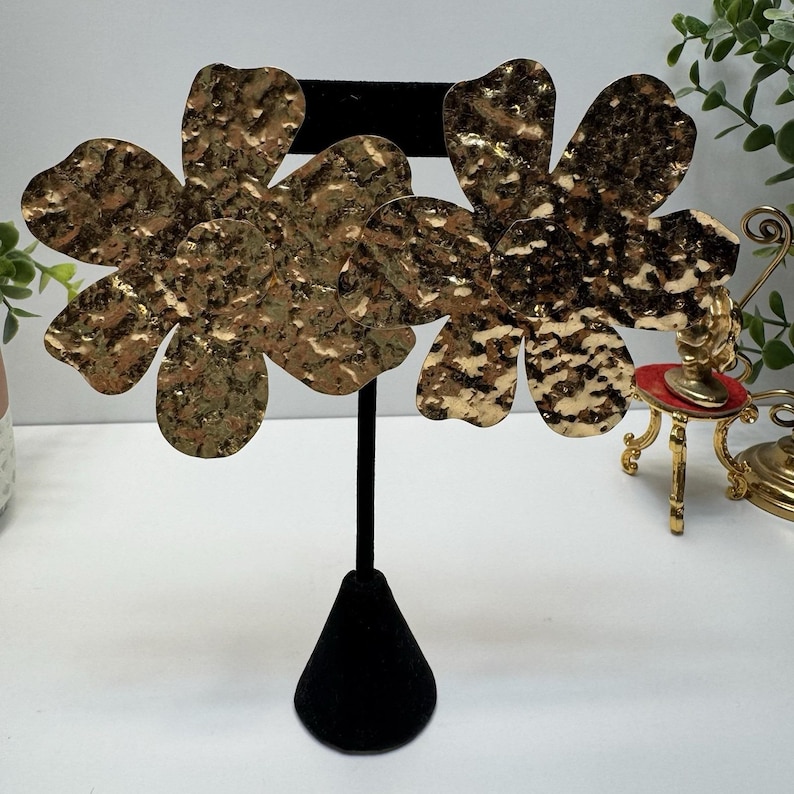 May include: Gold-toned, hammered metal flower earrings displayed on a black velvet jewelry stand. The earrings have a textured surface and a multi-toned gold and brown color scheme. The floral design is prominent, with each earring featuring multiple petals.