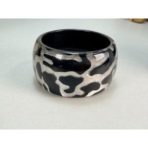 May include: A wide bangle bracelet featuring a high-contrast design with glossy black sections interspersed with irregular, organic-shaped metallic silver patterns that wrap around the entire circumference of the circular wrist accessory.