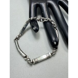 K&ouml;nnte beinhalten: Ein silbernes Armband mit einer Kette und rechteckigen und zylindrischen Gliedern. Das Armband hat dekorative ge&auml;tzte Muster auf den Gliedern und einen Verschluss. Das Armband wird auf einem schwarzen handf&ouml;rmigen Display pr&auml;sentiert.