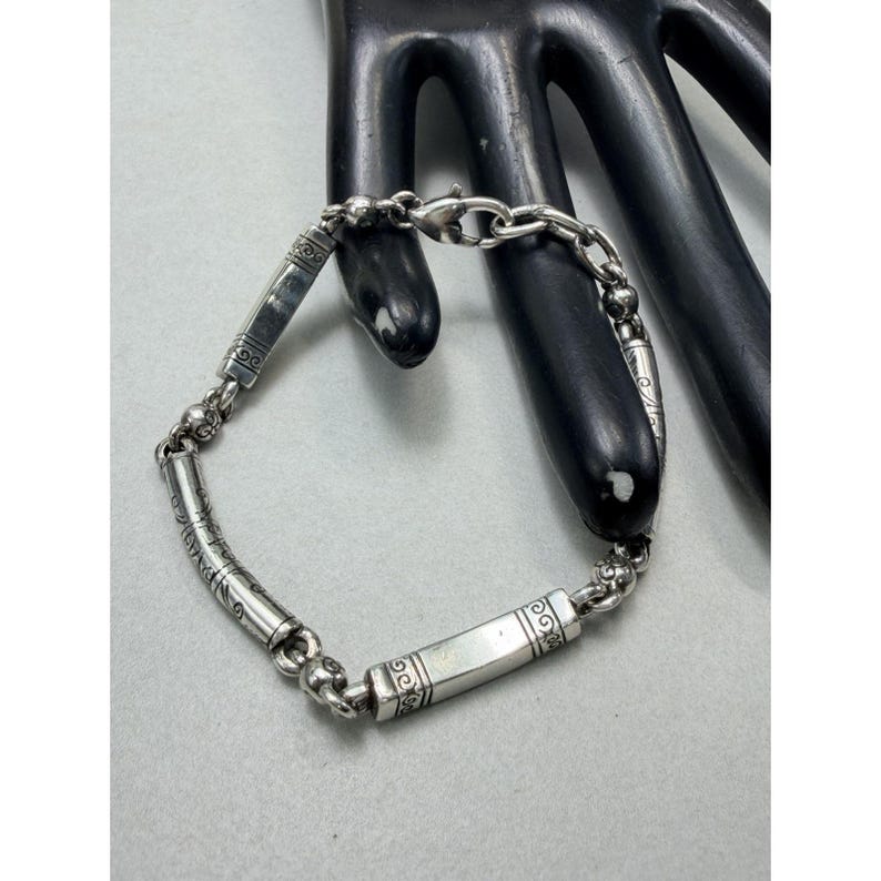 K&ouml;nnte beinhalten: Ein silbernes Armband mit einer Kette und rechteckigen und zylindrischen Gliedern. Das Armband hat dekorative ge&auml;tzte Muster auf den Gliedern und einen Verschluss. Das Armband wird auf einem schwarzen handf&ouml;rmigen Display pr&auml;sentiert.