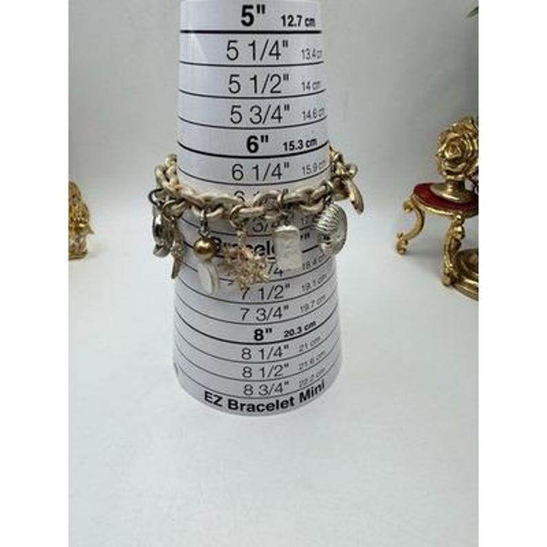 May include: A silver-toned charm bracelet with various charms, including shells, a heart, and a rectangular pendant. The bracelet is displayed on a white EZ Bracelet Mini measuring tool. The bracelet appears to be approximately 6 inches (15.3 cm) in diameter.