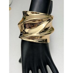 May include: A wide, polished gold-tone cuff bracelet featuring a highly reflective, smooth surface with deep, undulating, wave-like folds and curves that create a dynamic, sculptural appearance across the entire width of the metal band.