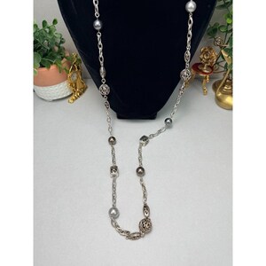 Może przedstawiać: Srebrny naszyjnik z łańcuszkiem, z szarymi koralikami, kwadratowymi zawieszkami i ozdobnymi, okrągłymi wisiorkami. Naszyjnik jest prezentowany na czarnym tle.