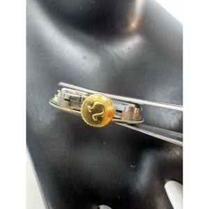 May include: A gold-colored button with the letter "S" in the center, attached to a silver-colored metal object. The button is round and has a textured surface. The metal object appears to be a clasp or fastener.