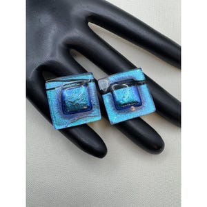 May include: A pair of square dichroic glass earrings. The earrings feature a vibrant blue and teal color scheme with a textured, iridescent finish. Each earring has a dark, rectangular accent. The earrings are displayed on a black surface.