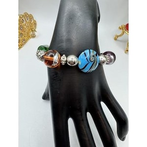 May include: A beaded bracelet featuring colorful glass beads in shades of green, orange, blue, and purple. The bracelet includes silver-toned metal beads and is displayed on a black hand-shaped display stand. The bracelet is a piece of jewelry.