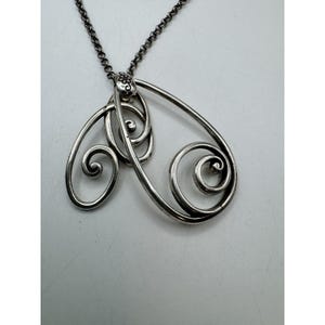 May include: A silver-toned pendant necklace featuring an openwork design with swirling, scroll-like shapes. The pendant is suspended from a delicate chain. The design includes a small floral detail at the top, adding a touch of elegance to the jewelry.