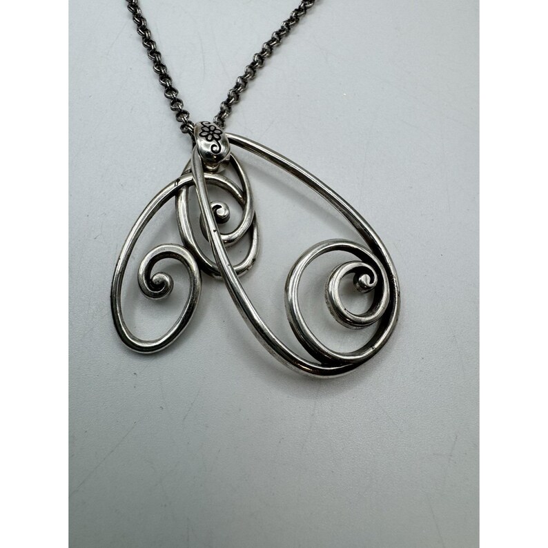May include: A silver-toned pendant necklace featuring an openwork design with swirling, scroll-like shapes. The pendant is suspended from a delicate chain. The design includes a small floral detail at the top, adding a touch of elegance to the jewelry.