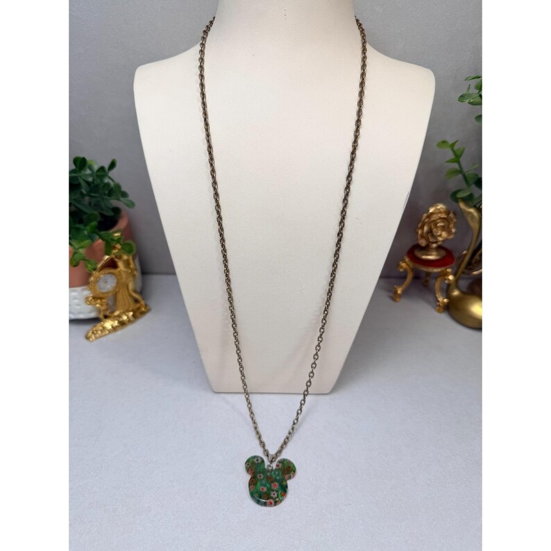 May include: A long, bronze-colored chain necklace with a pendant shaped like a mouse head. The pendant is green with a floral pattern in red, white, and pink. The necklace is displayed on a white mannequin.